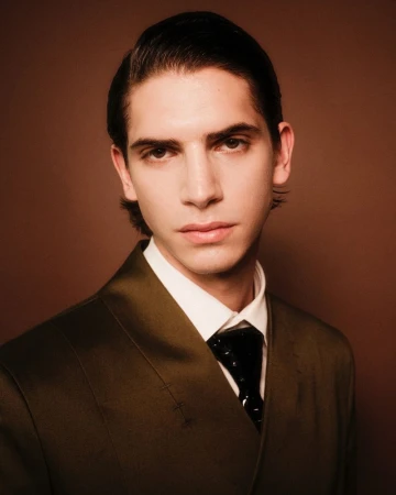 Headshot of a stylish adult male with dark hair, fair skin, and a serious expression, wearing a suit and tie against a brown backdrop.