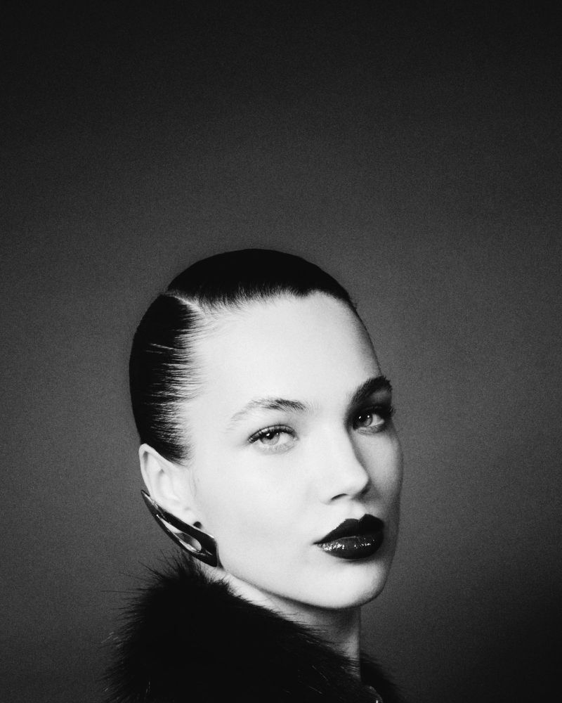 Monochrome portrait of a stylish adult woman with dark lipstick, slicked-back hair, and distinctive ear cuff jewelry against a dark backdrop.