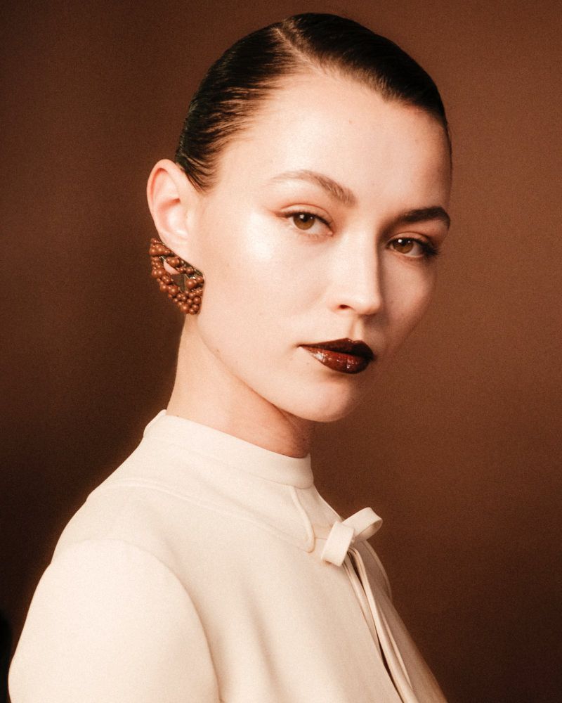 Close up portrait of a model with dark slicked hair wearing a cream high neck top and textured earrings against a brown backdrop