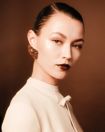 Close up portrait of a model with dark slicked hair wearing a cream high neck top and textured earrings against a brown backdrop