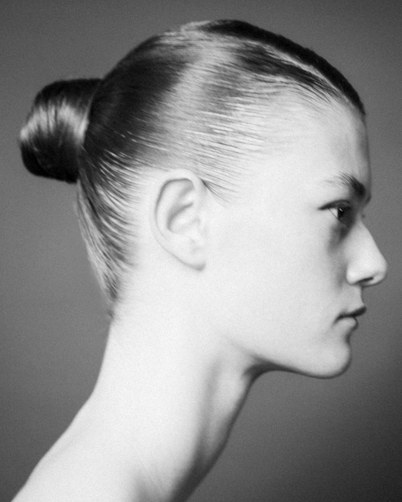 Black and white studio portrait of a woman with her hair in a bun against a gray background. She is facing to the right.