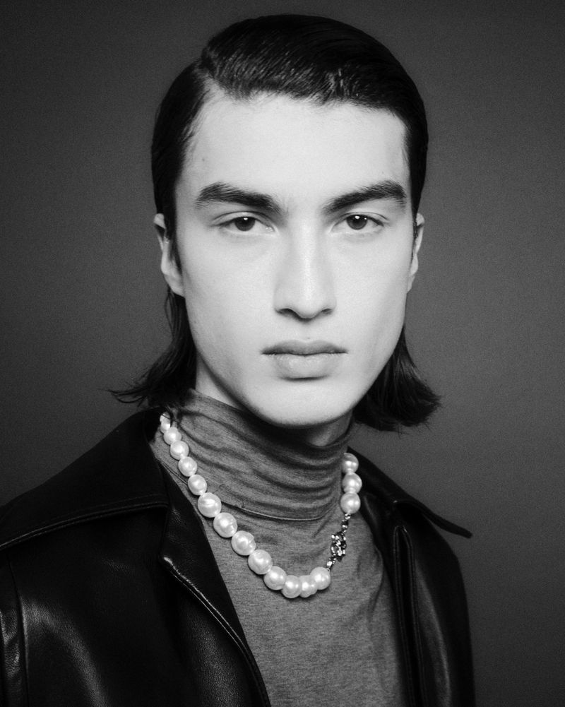 Black and white portrait of a young adult man with slicked back hair, wearing a gray turtleneck, leather jacket, and pearl necklace.