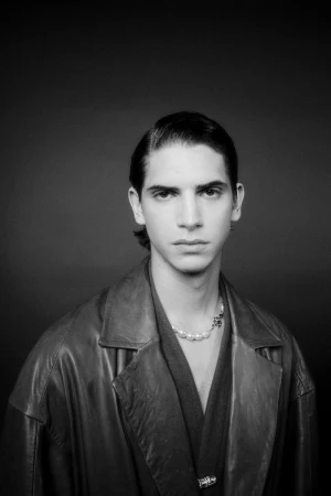 Monochrome portrait of a male model wearing a dark leather coat and pearl necklace against a dark background