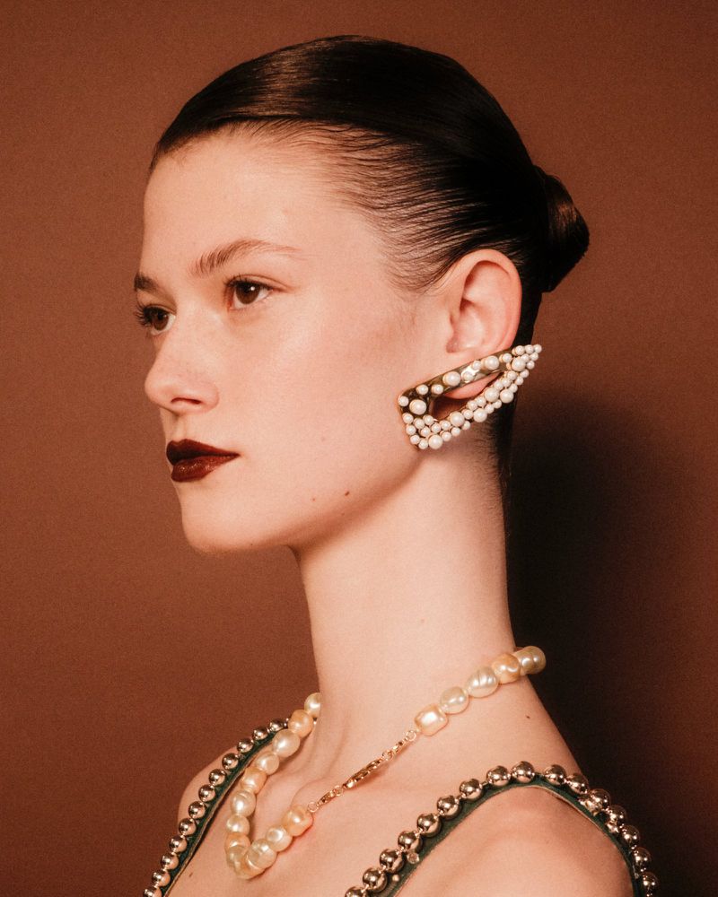 Profile view of an adult female fashion model with pearl-covered jewelry, dark lipstick, and sleek hair in an indoor setting.