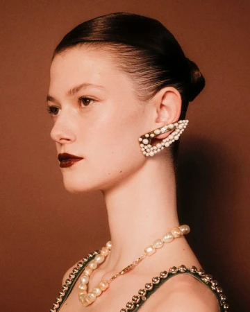 Profile view of an adult female fashion model with pearl-covered jewelry, dark lipstick, and sleek hair in an indoor setting.