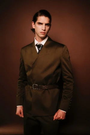 Model in olive green suit and tie with belted jacket posing against solid brown backdrop for Georges Hobeika F/W 25 Backstages.