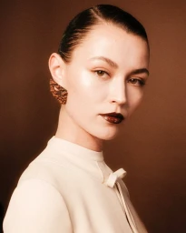 Close up portrait of a model with dark slicked hair wearing a cream high neck top and textured earrings against a brown backdrop