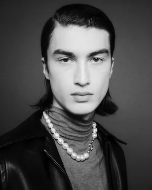 Portrait of a male model wearing a pearl necklace, turtleneck, and dark jacket against a plain background
