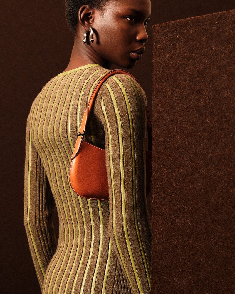 Elegant woman in a striped dress and sculptural earrings poses against a brown backdrop with a satchel purse on her shoulder.