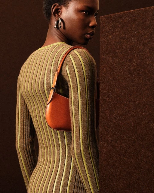 Elegant woman in a striped dress and sculptural earrings poses against a brown backdrop with a satchel purse on her shoulder.
