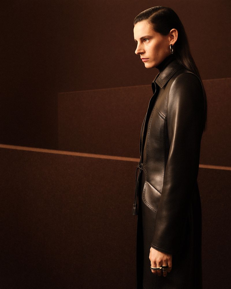 Model in dark leather coat with slicked-back dark hair posing against a warm brown architectural background.