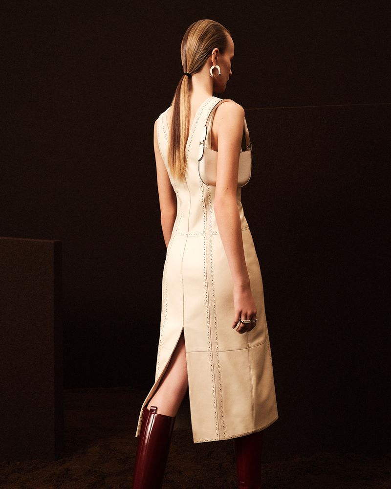 Back view of a woman in a sleeveless cream dress and burgundy boots with a shoulder bag in a studio setting against a dark background.