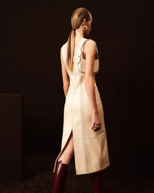 Back view of a woman in a sleeveless cream dress and burgundy boots with a shoulder bag in a studio setting against a dark background.