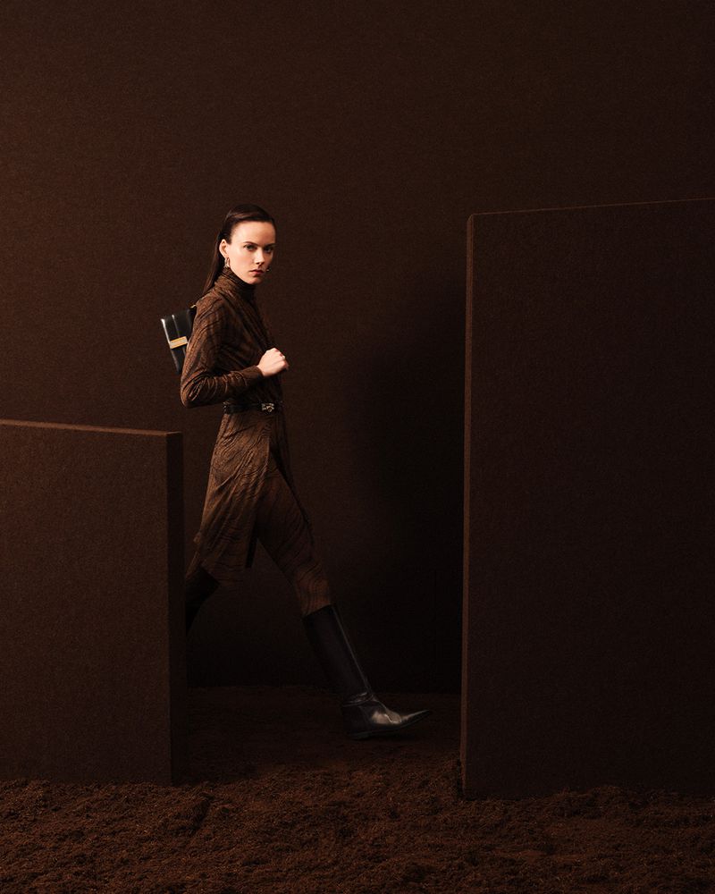 Fashion shot of a woman in a brown patterned coat dress, wearing black boots, and a small black bag, walking on textured ground.