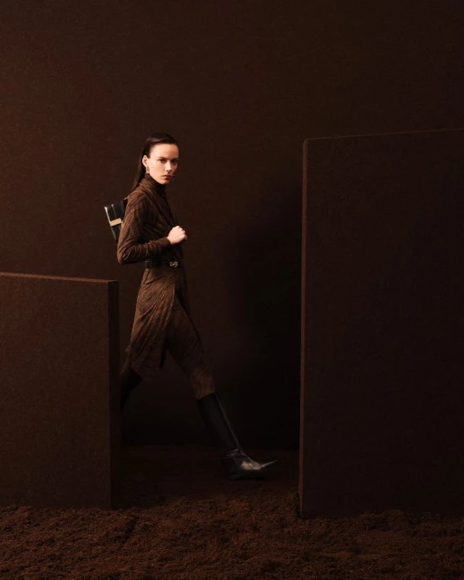 Fashion shot of a woman in a brown patterned coat dress, wearing black boots, and a small black bag, walking on textured ground.