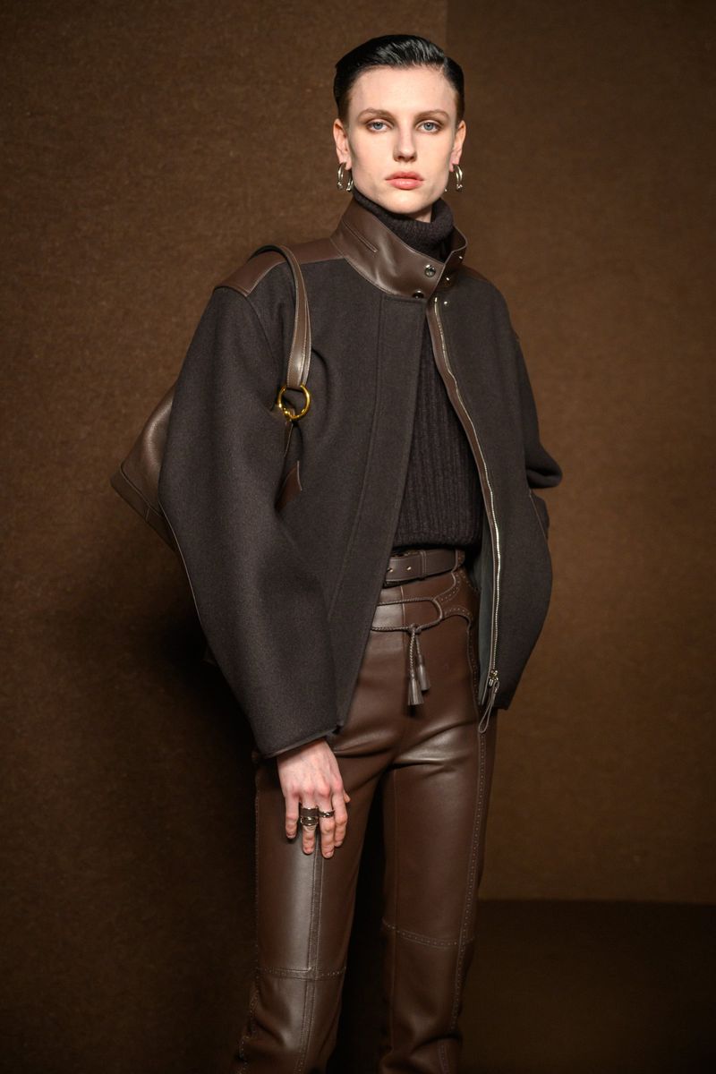 Portrait of a stylish woman in a black jacket and brown leather pants, carrying a brown leather shoulder bag against a brown background.