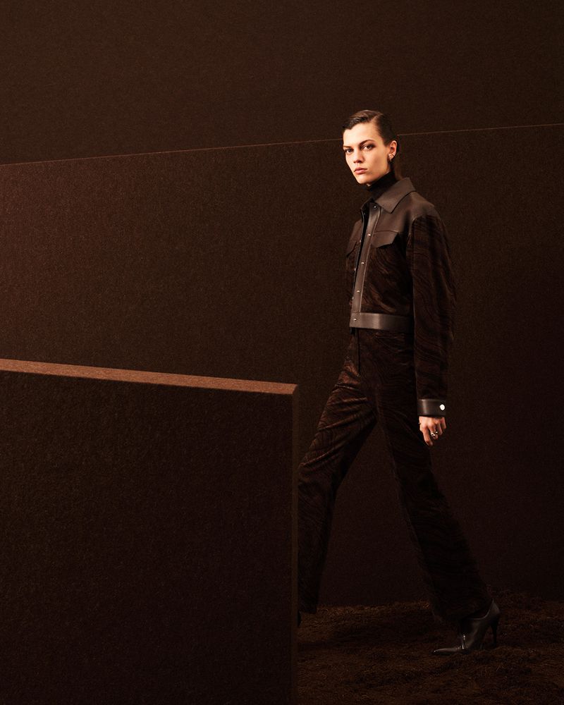 Model wearing dark brown jacket and trousers walking on a textured brown set design for Hermes Fall/Winter 2025.