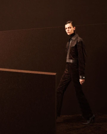 Model wearing dark brown jacket and trousers walking on a textured brown set design for Hermes Fall/Winter 2025.