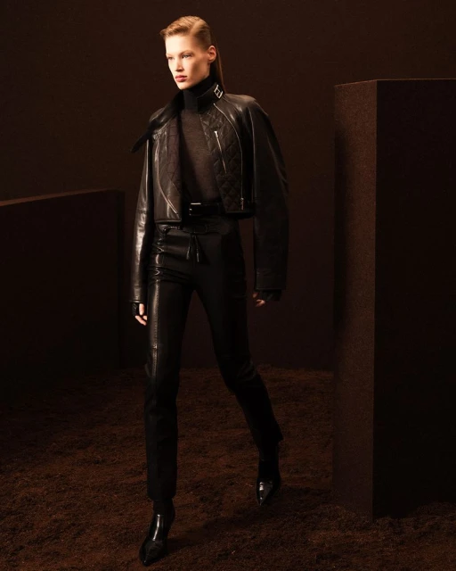 A female fashion model poses in a black leather jacket and pants in a studio with a dark brown textured floor.