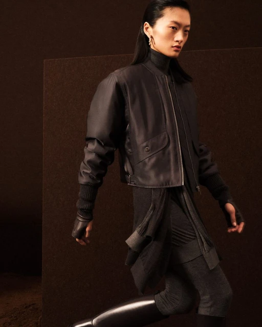 Fashionable Asian woman wearing a black bomber jacket and leggings, posing in a studio with a dark brown backdrop for a fashion editorial.