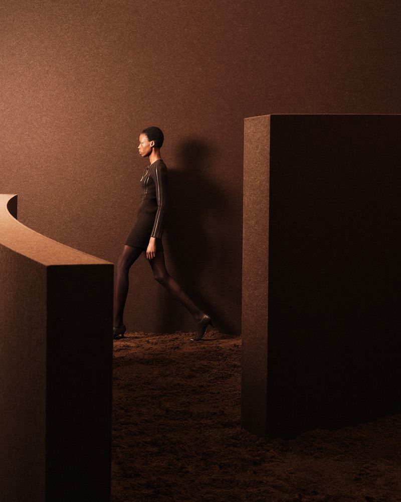 Woman in black dress posing in a studio with brown monochromatic backdrop and abstract set. Fashion editorial shot with geometric set design.