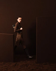 Model in brown turtleneck dress and knee-high boots walking between dark partitions