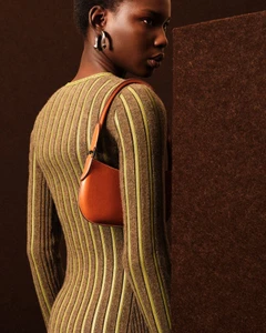 Model wearing a ribbed knit dress and carrying an orange leather shoulder bag against a dark studio backdrop