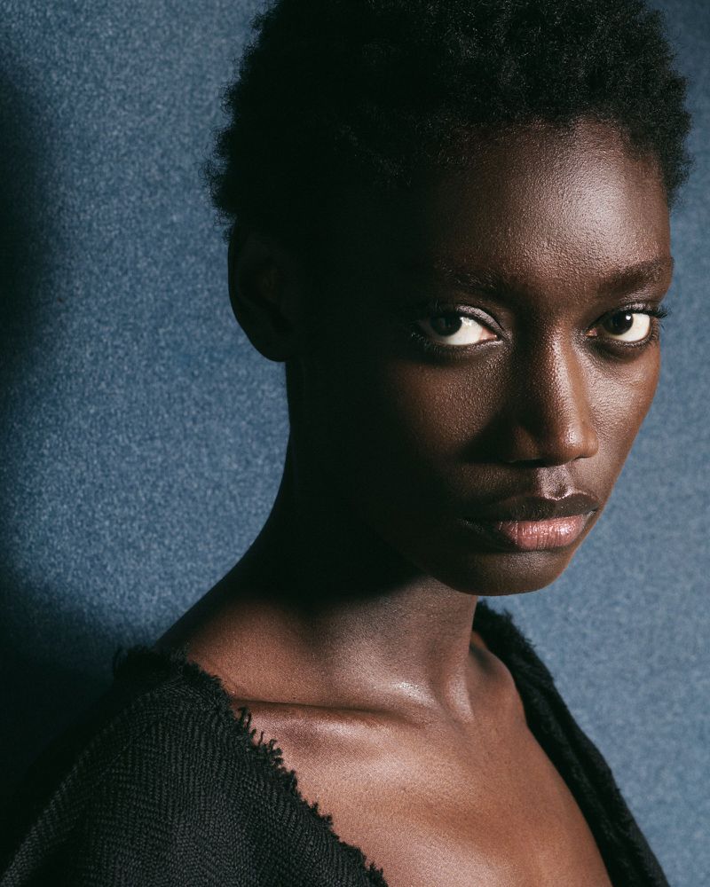 Close-up studio portrait of a dark-skinned adult female with short hair, wearing a black woven top, against a textured blue background.