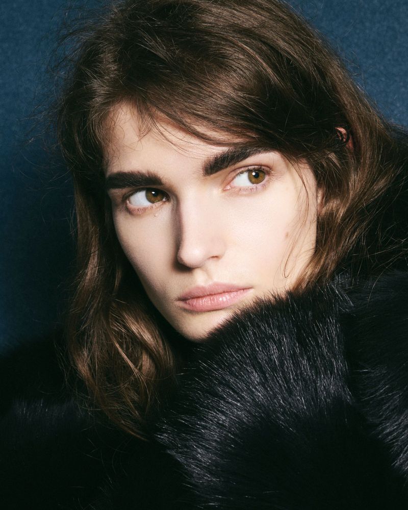 Close-up studio portrait of a young adult woman with brown hair and eyes, wearing a black fur coat, gazing off-camera against a dark background.