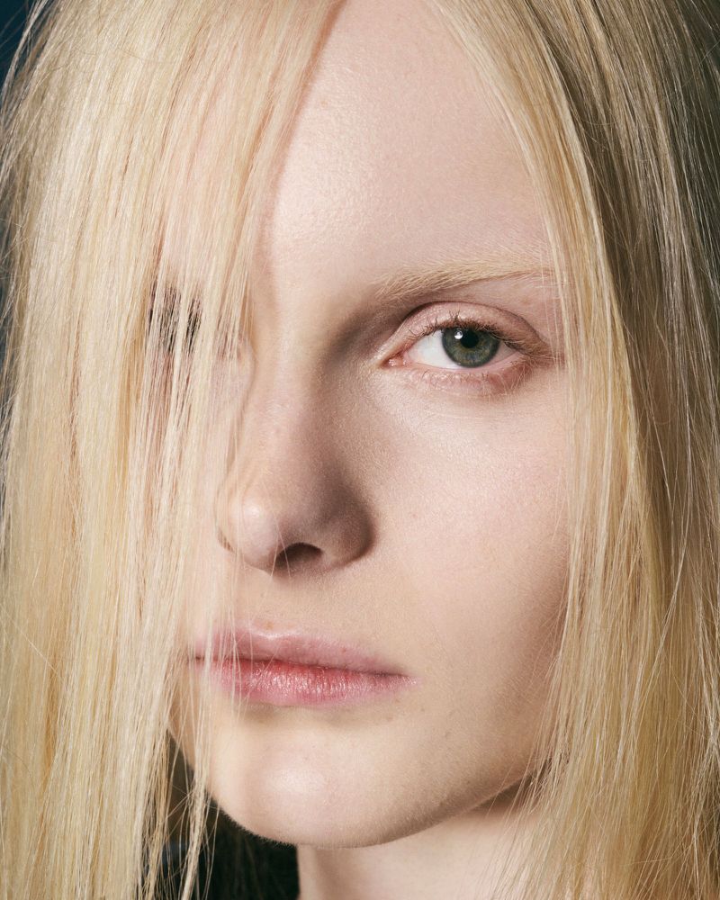 Close-up portrait of a blonde-haired young adult woman with light skin and green eyes, one eye visible and hair covering part of the face.