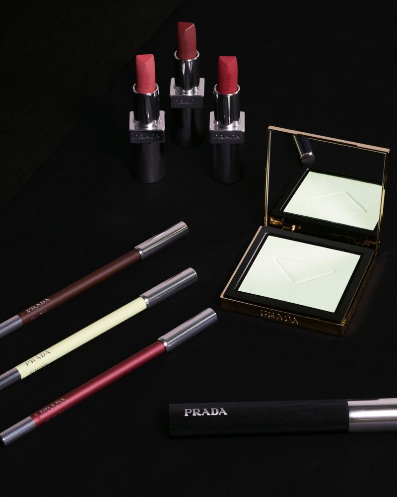 Cosmetic still life features a collection of Prada lipsticks, eyeliners, powder compact, and mascara on a black background.