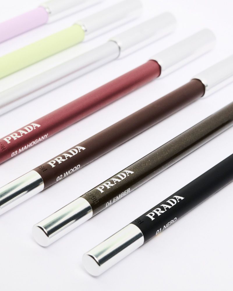 Close-up of Prada eyeliner pencils in assorted shades including mahogany, wood, ember, and nero arranged diagonally on a white surface.