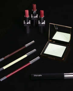 Prada Beauty makeup products including three lipsticks, three cosmetic pencils, a compact powder with mirror, and a tube against a black background
