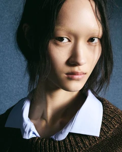 Close-up portrait of a model with dark hair wearing a light blue collared shirt and brown knitted sweater against a textured blue background.