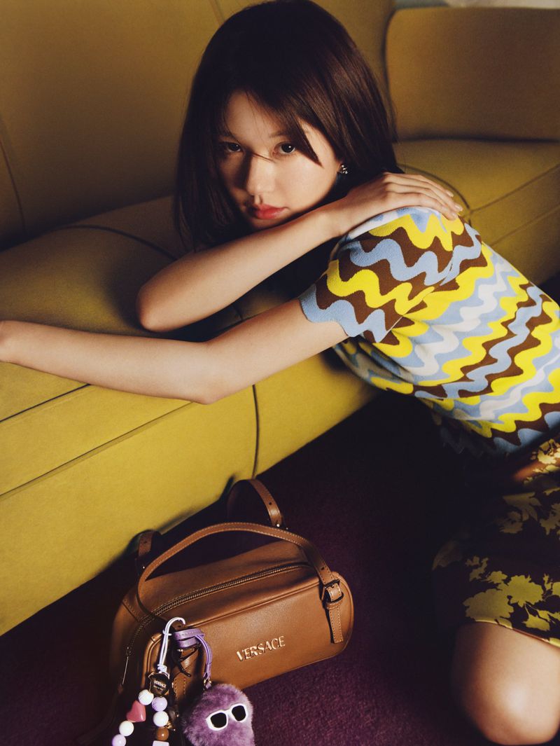 A stylish young woman with dark hair poses indoors on a yellow sofa, wearing a patterned top and a brown Versace handbag with a purple charm.