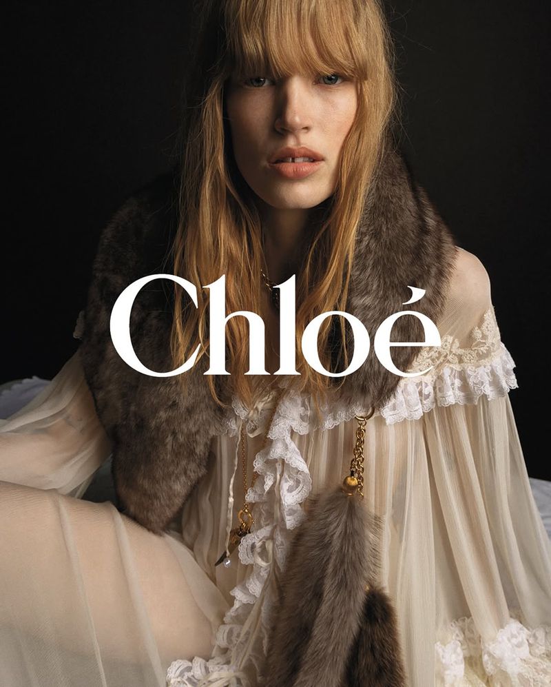 Close-up of a fair-skinned, blonde woman with bangs, wearing a sheer white dress and fur stole, posing in front of a black backdrop.