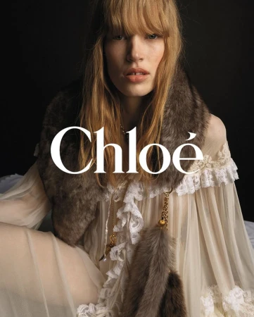 Close-up of a fair-skinned, blonde woman with bangs, wearing a sheer white dress and fur stole, posing in front of a black backdrop.