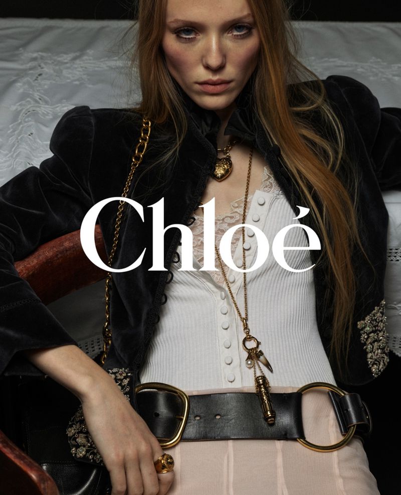 Fashion shot of a young woman with long reddish blonde hair wearing a black jacket and gold chain with Chloe brand logo.
