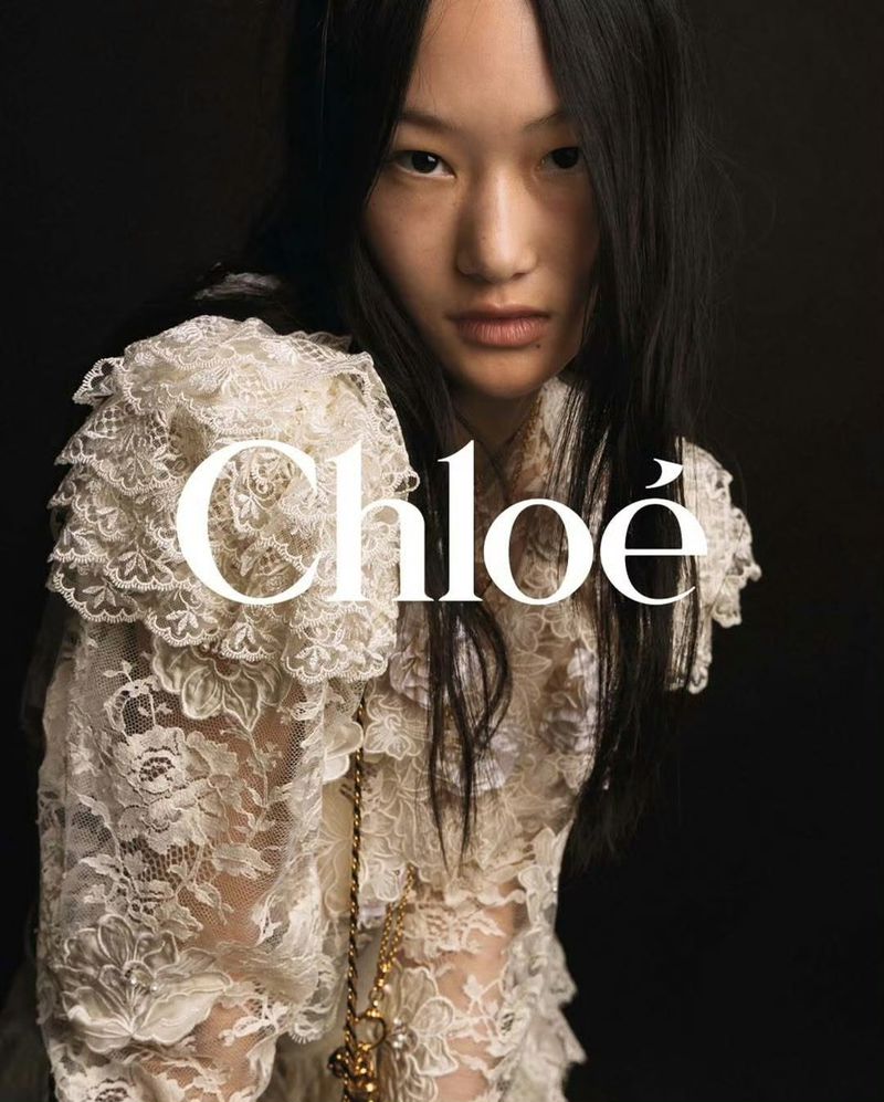 Portrait of a young Asian woman wearing a frilly lace top with the 'Chloe' logo. A golden pendant hangs on a chain. Dark studio background.