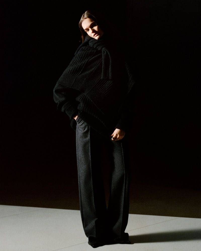 Fashion shot of an adult woman in a black knit turtleneck and dark grey slacks, standing in a photo studio with dark background.