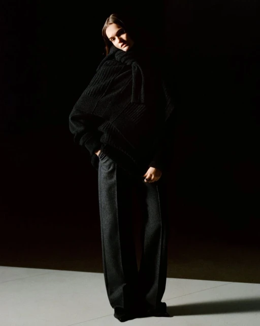 Fashion shot of an adult woman in a black knit turtleneck and dark grey slacks, standing in a photo studio with dark background.