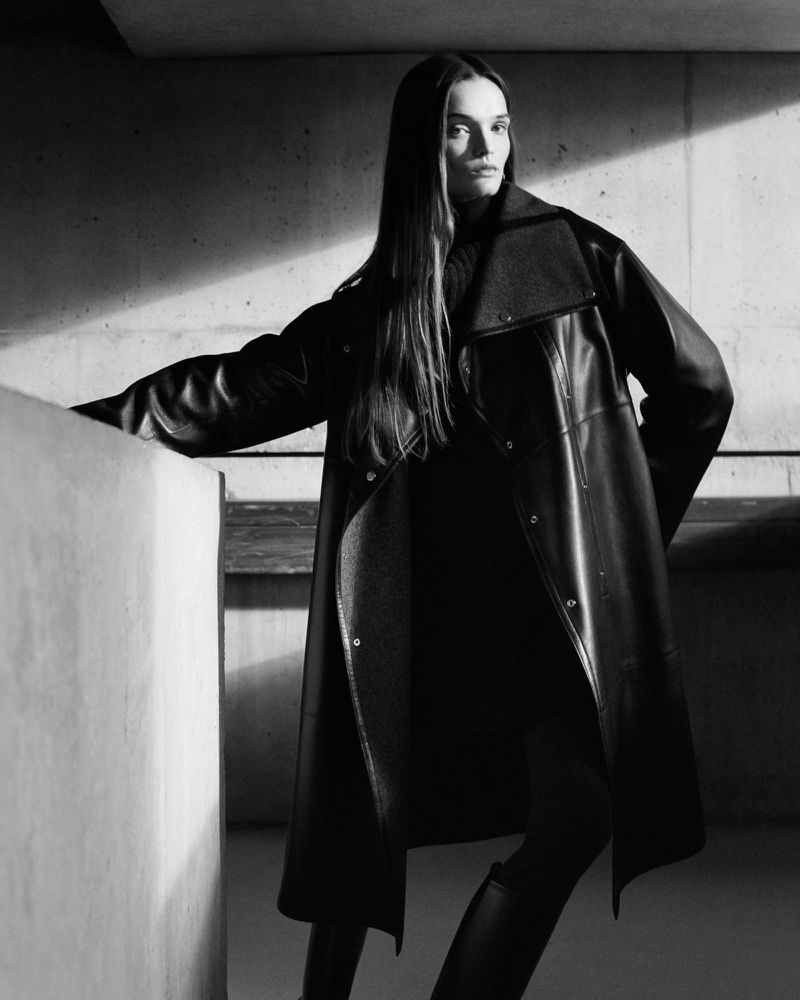 Monochrome fashion shot: woman with long hair poses in an oversized black leather trench coat in front of a concrete wall indoors.