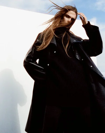Model Lulu Tenney in a black coat with long hair blowing outdoors against a white wall.