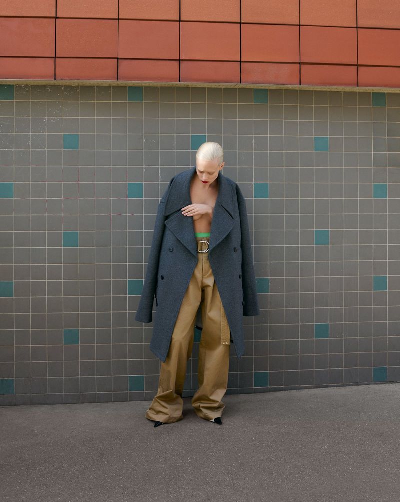A fashionable adult woman with short bleached hair poses outdoors in an oversized coat and khaki pants against a tiled wall.