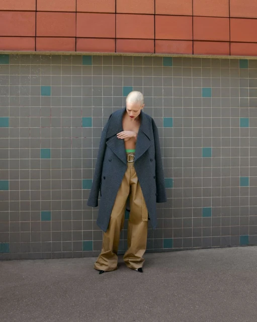 A fashionable adult woman with short bleached hair poses outdoors in an oversized coat and khaki pants against a tiled wall.