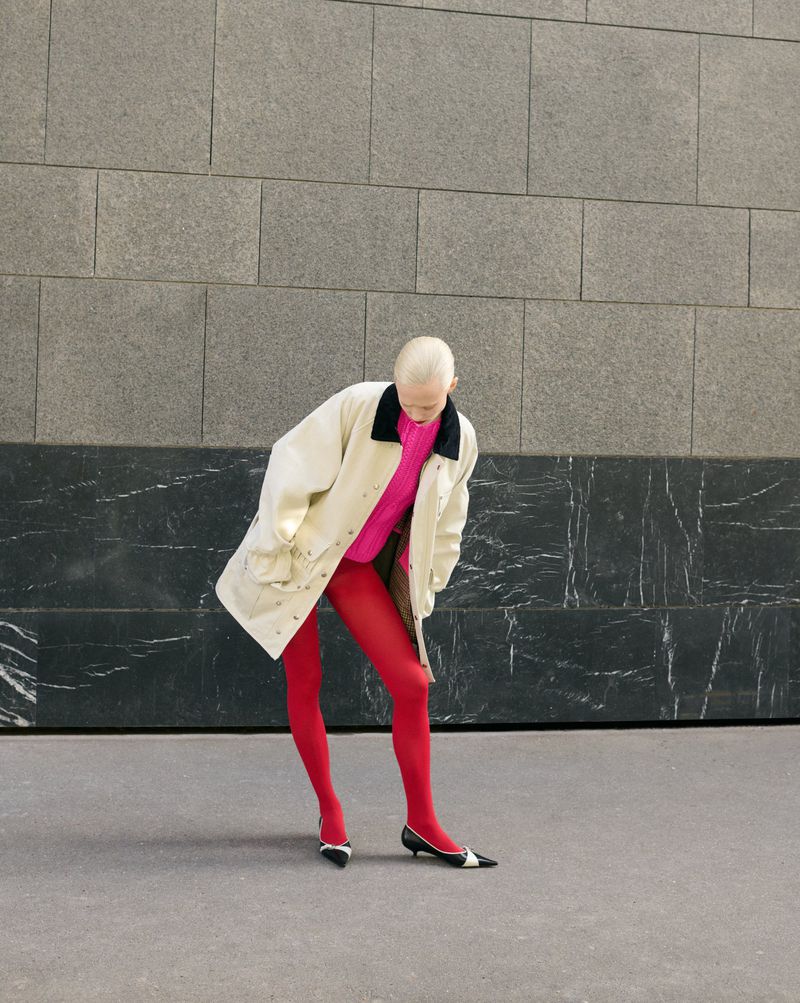Woman in bright pink sweater and red tights posing against an abstract wall in a beige coat and black and white shoes. 