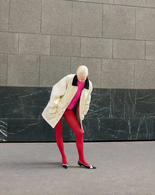 Woman in bright pink sweater and red tights posing against an abstract wall in a beige coat and black and white shoes. 