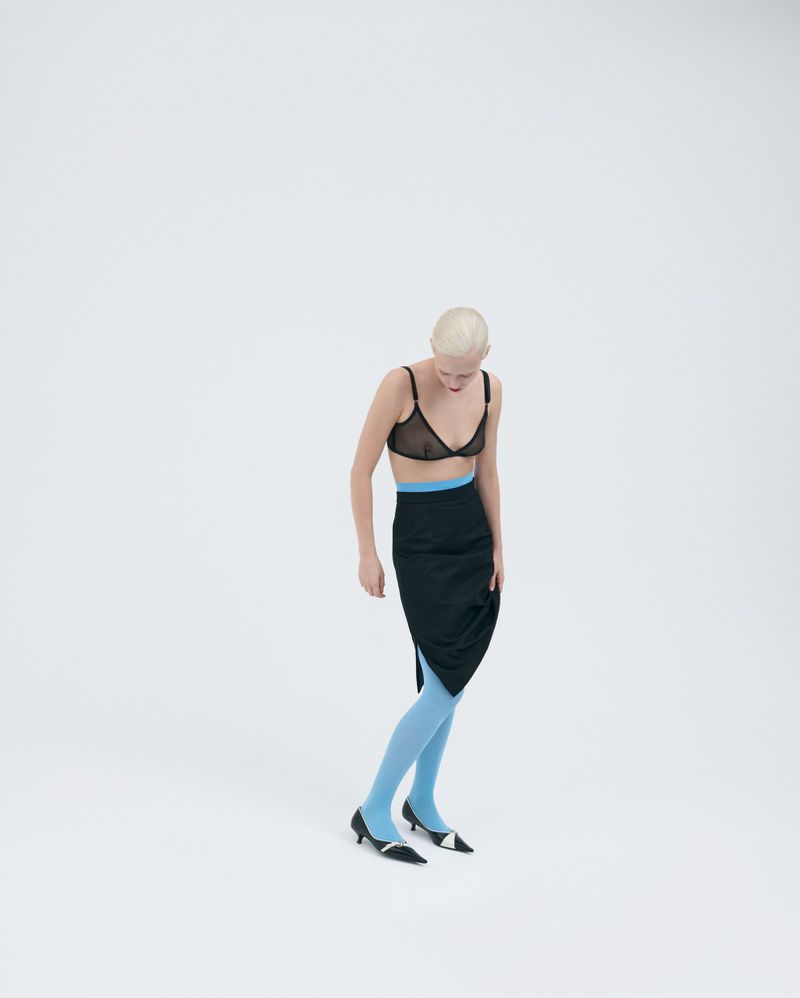 A slender bald woman in a black bra, skirt, and blue hosiery poses in a minimalist fashion shot in a white studio setting. 