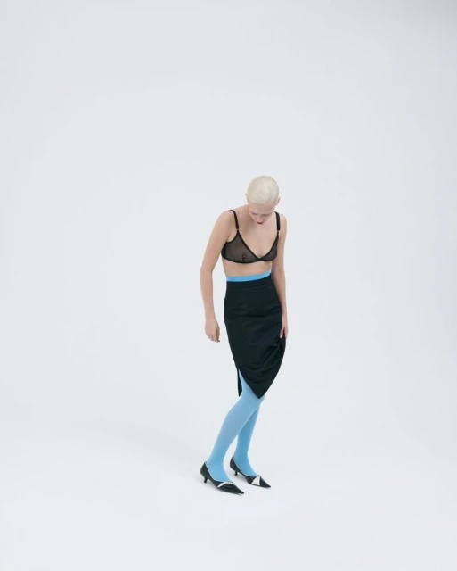 A slender bald woman in a black bra, skirt, and blue hosiery poses in a minimalist fashion shot in a white studio setting. 