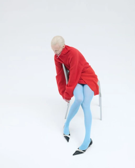 A fair-skinned person sits on a metal chair in a photo studio wearing a red sweater, blue tights, and black heels against a white backdrop.